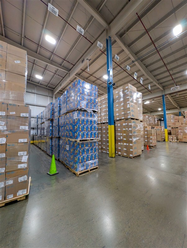 Clean warehouse aisle with organized pallets of finished goods, marked floor zones, and pest monitoring stations visible on wall